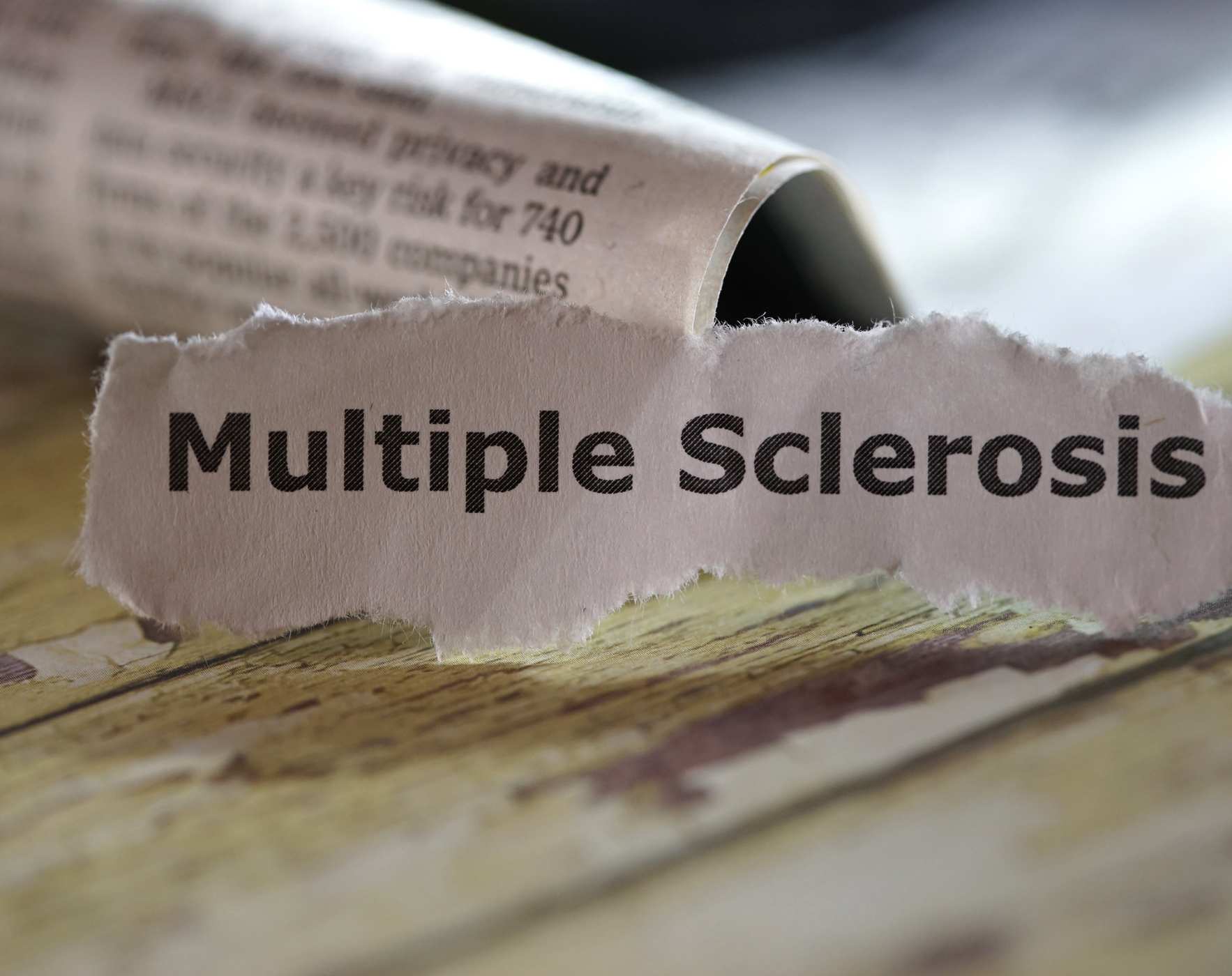 Close-up of a torn newspaper clipping with the words Multiple Sclerosis printed in bold black text on a wooden surface.